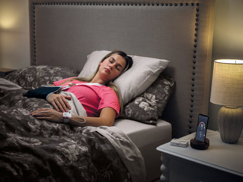 Patient laying in bed wearing the safetynet alert monitor with home medical hub and phone displaying Masimo Halo App on nightstand. 